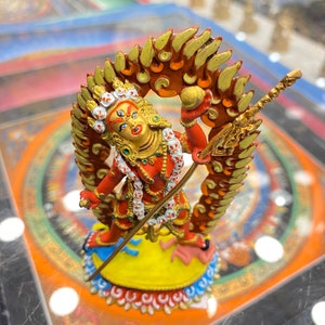 May include: A red and gold statue of a deity with a fierce expression, holding a spear and standing on a yellow base with a blue and white border. The deity is surrounded by a red and gold halo with flames.