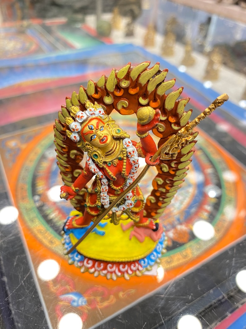 May include: A red and gold statue of a deity with a fierce expression, holding a spear and standing on a yellow base with a blue and white border. The deity is surrounded by a red and gold halo with flames.