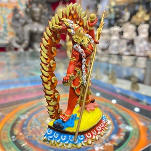 May include: A red and gold figurine of a deity with a flaming halo, holding a staff and standing on a blue and yellow base. The figurine is on a colorful mandala-like surface.