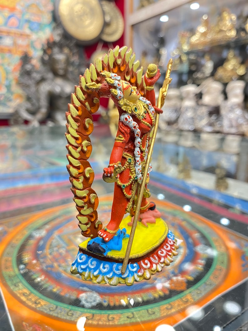 May include: A red and gold figurine of a deity with a flaming halo, holding a staff and standing on a blue and yellow base. The figurine is on a colorful mandala-like surface.