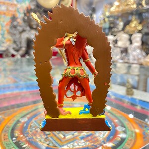 May include: A red and gold statue of a deity with a fierce expression, standing on a yellow base with a blue and gold mandala design. The statue is surrounded by a gold halo with a flame-like design.