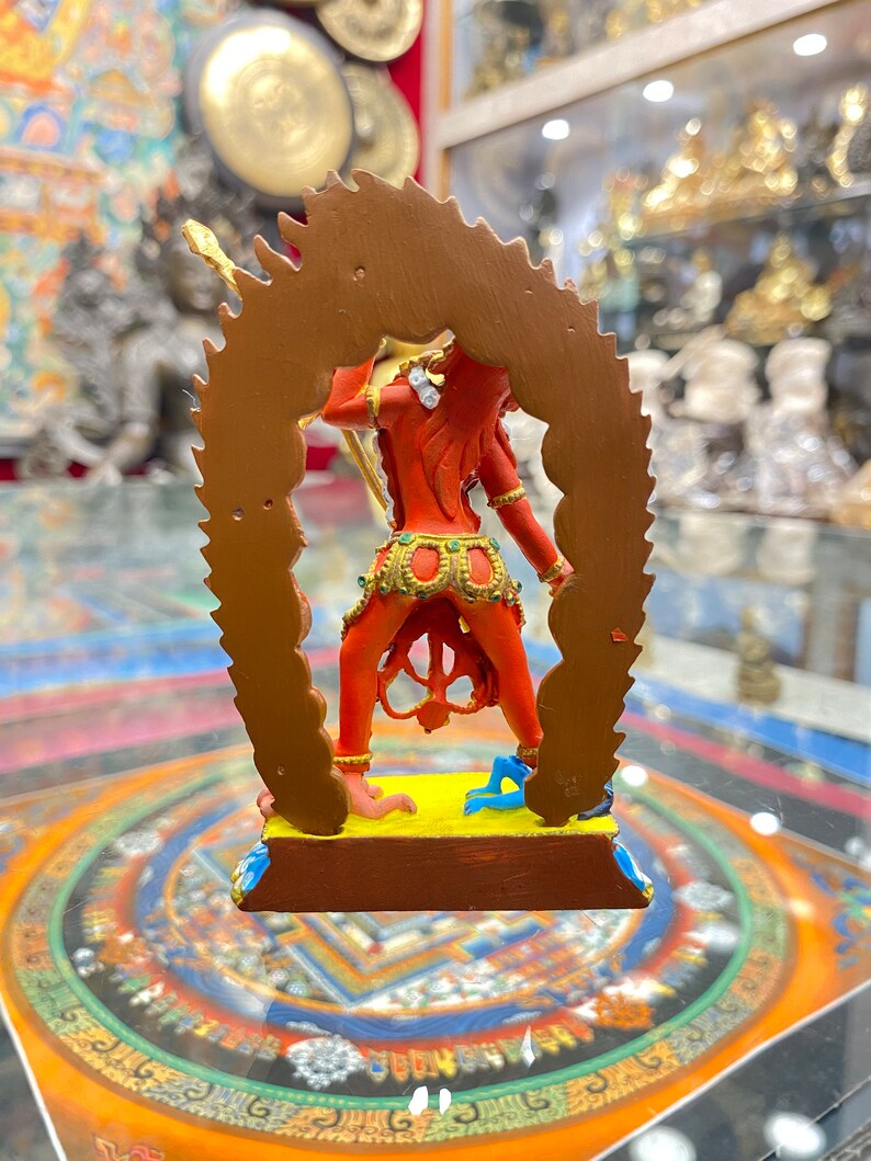 May include: A red and gold statue of a deity with a fierce expression, standing on a yellow base with a blue and gold mandala design. The statue is surrounded by a gold halo with a flame-like design.