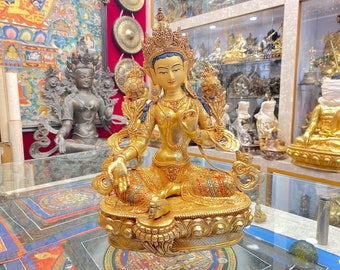 May include: A golden statue of a deity with a crown and intricate details. The statue is sitting in a meditative pose with hands in a mudra gesture. The statue is adorned with colorful jewels and is set against a backdrop of a shop display.