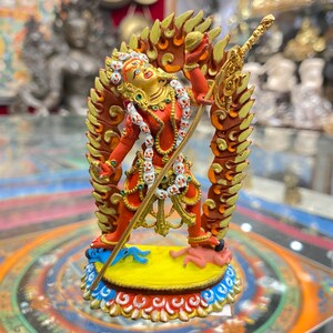 May include: A red and gold figurine of a deity with a flaming halo, holding a sword and a sphere. The deity is standing on a yellow base with blue and red accents. The figurine is surrounded by a circular mandala design.