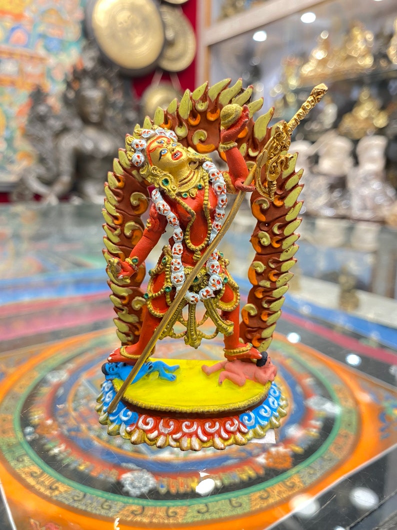 May include: A red and gold figurine of a deity with a flaming halo, holding a sword and a sphere. The deity is standing on a yellow base with blue and red accents. The figurine is surrounded by a circular mandala design.