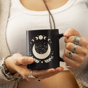May include: Black ceramic mug with a white moon and floral design. The mug has a black handle and a white crescent moon with a floral design inside.