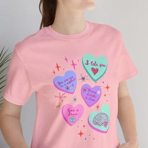 Puede incluir: Camiseta rosa con gr&aacute;ficos en forma de coraz&oacute;n en varios colores, incluyendo azul, verde y rosa. Los corazones presentan texto e ilustraciones relacionadas con el cerebro, con frases como "I love you" y "Time is brain".