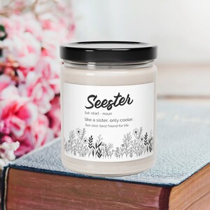 May include: A white candle jar with a black lid and a label that reads "Seester" with the definition "like a sister, only cooler. See also: best friend for life". The candle jar is sitting on a closed book.