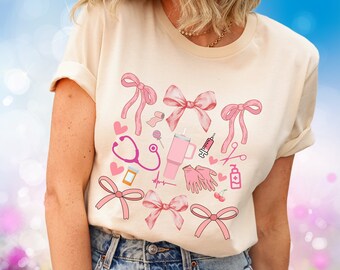 May include: A light beige t-shirt with a pink and white design featuring a stethoscope, a heart rate monitor, a syringe, a bandage, a lollipop, a cup, a pair of scissors, a pair of gloves, and a cherry. The design is surrounded by pink bows.