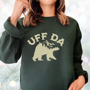 May include: Dark green sweatshirt with the text "UFF DA" above a bear silhouette with a mountain and tree design. The sweatshirt is a solid color with long sleeves and a crew neck.