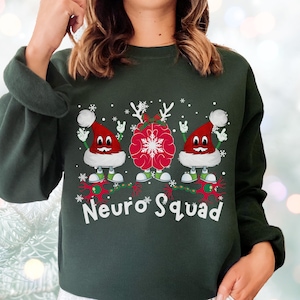 May include: Dark green sweatshirt with a festive design. The graphic features two cartoon brains wearing Santa hats and a central brain with snowflake antlers. The text "Neuro Squad" is below the design. Snowflakes are scattered around the design.