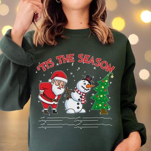 May include: Dark green sweatshirt with a festive graphic. The design features Santa Claus, a snowman, and a Christmas tree. The text "'Tis the Season" is printed across the top in red. The sweatshirt is a comfortable, casual style.