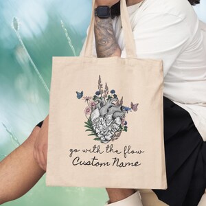 May include: A beige tote bag with a graphic of a human heart surrounded by flowers and butterflies. The text "go with the flow Custom Name" is printed below the graphic.