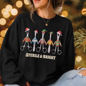 May include: A black crewneck sweatshirt featuring a graphic of five surgical hemostats wearing colorful Christmas sweaters and Santa or elf hats. The text "STERILE & BRIGHT" is printed in a cream-colored font below the illustrated medical tools.