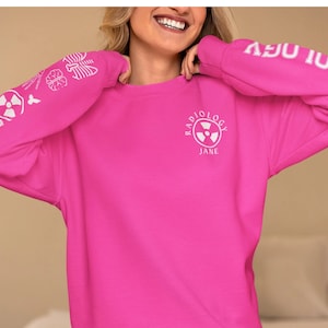May include: A pink long-sleeved sweatshirt with a white graphic on the front that says "RADIOLOGY JANE" with a radiation symbol. The sleeves have white graphics of medical symbols.