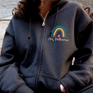May include: Black zippered hoodie with a rainbow and apple design and the text "Mrs. Patterson" on the front.