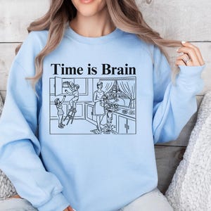 May include: Light blue sweatshirt with the phrase "Time is Brain" above a black and white illustration of a chaotic kitchen scene. The illustration depicts a woman and children in a kitchen.