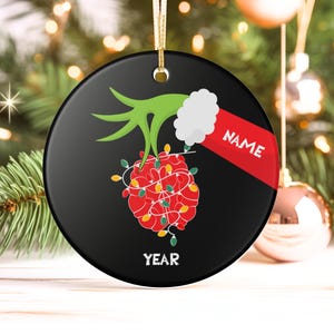 May include: Black round Christmas ornament with a green hand holding a red ball wrapped in Christmas lights. The ornament has the word "NAME" on a red banner and the word "YEAR" at the bottom.