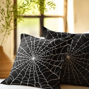 May include: Two black throw pillows with a white spiderweb design. The pillows are on a white surface.