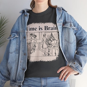 May include: Dark gray t-shirt with the text "Time is Brain" above a black and white illustration of a kitchen scene. The illustration depicts people in a kitchen. The person is wearing a light blue denim jacket.