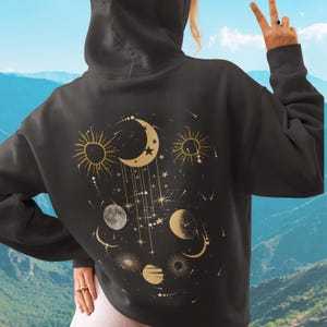 May include: Black hoodie with a celestial design on the back. The design features gold suns, moons, stars, and other celestial elements. The hoodie has a hood and a relaxed fit, suitable for casual wear.