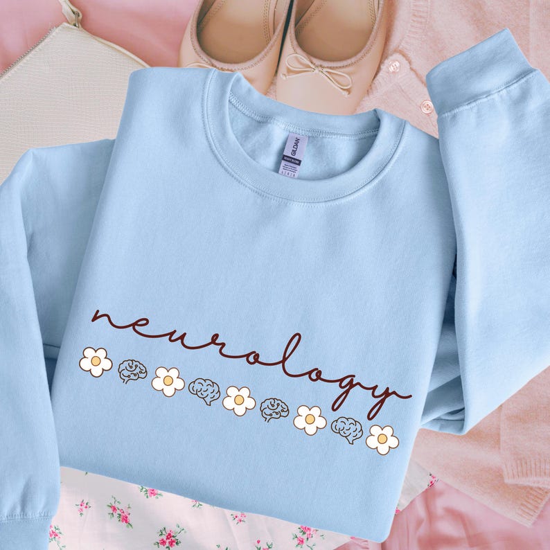 Puede incluir: Sudadera azul claro con cuello redondo con la palabra "neurology" en fuente manuscrita. La palabra est&aacute; decorada con flores blancas e ilustraciones de cerebros. La sudadera est&aacute; hecha de un material suave.