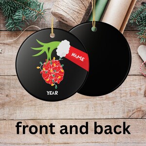 May include: Black round Christmas ornament with a festive design. The front features a red and yellow light-covered ornament with a green hand and white cuff. The word "NAME" is on a red banner. The word "YEAR" is at the bottom.