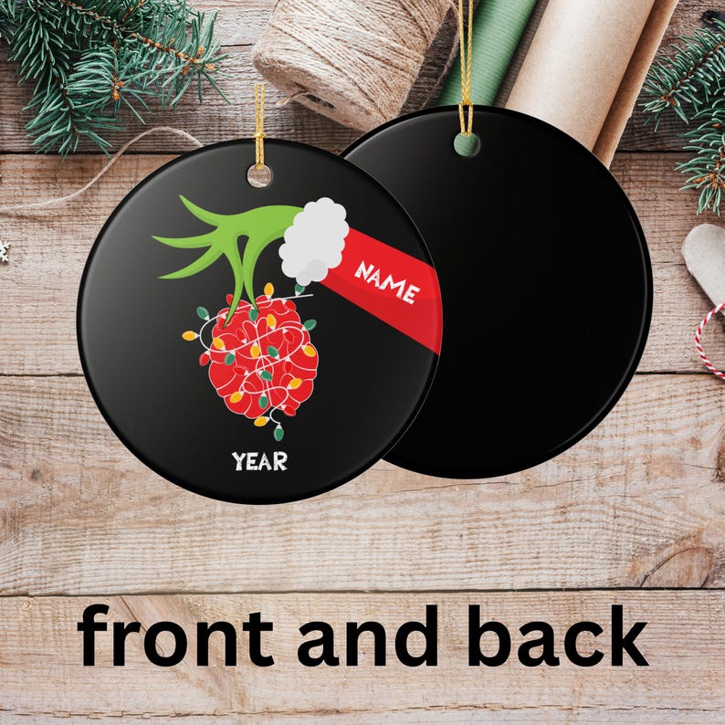 May include: Black round Christmas ornament with a festive design. The front features a red and yellow light-covered ornament with a green hand and white cuff. The word "NAME" is on a red banner. The word "YEAR" is at the bottom.