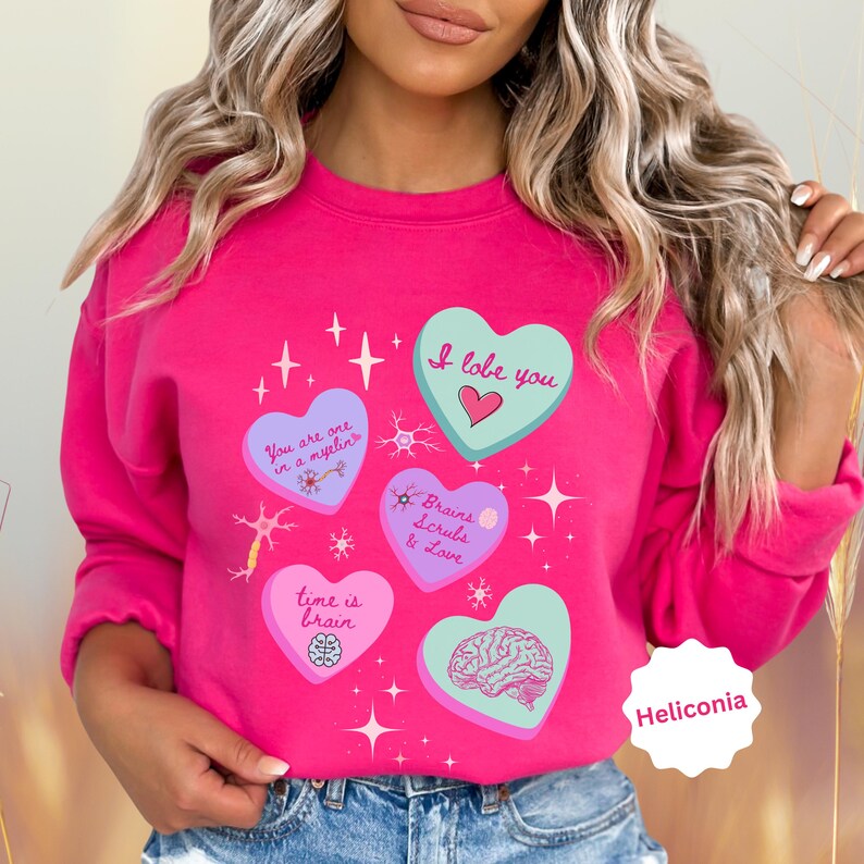 May include: A bright pink sweatshirt with several heart-shaped graphics. The hearts feature phrases like "I love you," "Brains, Scrubs & Love," and "Time is brain." The design includes brain illustrations and star accents. The word "Heliconia" is also visible.