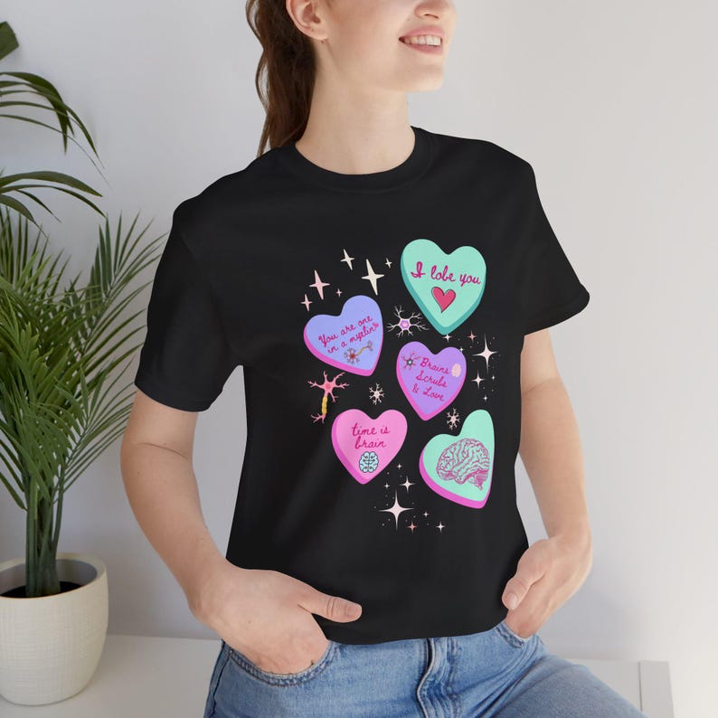 Puede incluir: Camiseta negra con un dise&ntilde;o de caramelos en forma de coraz&oacute;n de colores, cada uno con una frase diferente como "I lobe you" y "time is brain". El dise&ntilde;o tambi&eacute;n incluye un gr&aacute;fico de cerebro y acentos de estrellas.