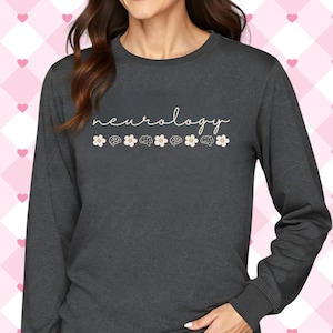 Puede incluir: Camiseta de manga larga gris oscuro con la palabra "neurology" en cursiva. Debajo, seis ilustraciones de cerebros se alternan con flores blancas con centros beige. La camiseta tiene cuello redondo y mangas largas.