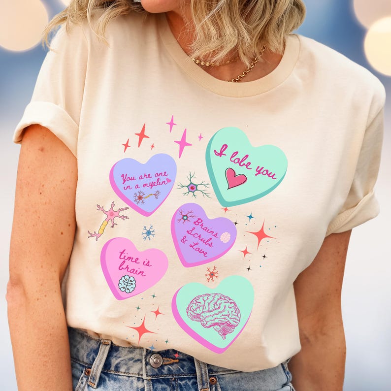 May include: Cream-colored t-shirt featuring heart-shaped graphics with medical-themed text and illustrations. The hearts are in shades of pink and teal, with phrases like "I lobe you" and "Brains Scrubs & Love". The shirt has rolled-up sleeves.