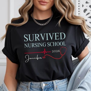 May include: Black t-shirt with the text "SURVIVED NURSING SCHOOL 2026" in light blue. A red heartbeat line with a stethoscope design and the name "Jennifer" in cursive. The shirt has rolled-up sleeves.