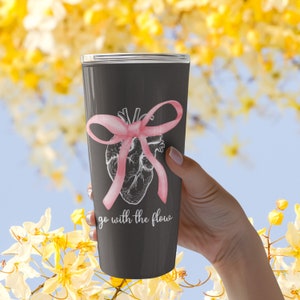 May include: A black and gray stainless steel tumbler with a pink ribbon tied around a stylized anatomical heart. The tumbler has the text "go with the flow" printed on the front.