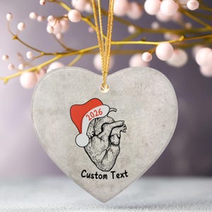 May include: Heart-shaped ornament with a gray, textured surface. The ornament features a detailed anatomical heart illustration wearing a red Santa hat with "2026" on it. The words "Custom Text" are printed below the heart.