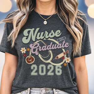 May include: Dark gray t-shirt with the words "Nurse Graduate 2026" in a retro font. The design includes a stethoscope, flowers, and a syringe. The shirt is a great gift for a nursing school graduate.