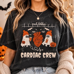 May include: Dark gray t-shirt with a Halloween-themed design. The shirt features the text "quit fibbin'" above a heart and two ghost characters wearing orange hats and holding drinks. The words "CARDIAC CREW" are printed below.