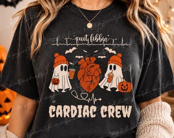 May include: Dark gray t-shirt with a Halloween-themed design. The shirt features the text "quit fibbin'" above a heart and two ghost characters wearing orange hats and holding drinks. The words "CARDIAC CREW" are printed below.
