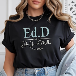 May include: Black t-shirt with the text "Ed.D Dr. Jane Mills Est. 2026" in light blue and white. The shirt has rolled-up sleeves and is made of a soft material. The shirt is a great gift for a doctor of education.