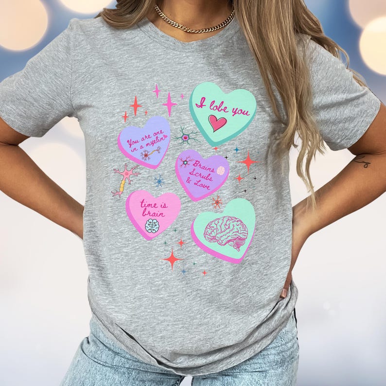 May include: A gray t-shirt featuring several heart-shaped designs with medical-themed phrases and illustrations. The hearts are in various colors, including pink, blue, and green, with phrases like "I lobe you" and "Brains Scrubs & Love".