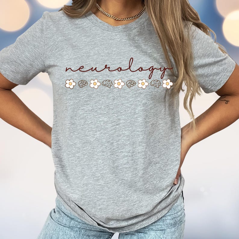 Puede incluir: Camiseta gris claro con la palabra "neurology" en escritura burdeos. Debajo, una fila de flores blancas y dibujos de cerebros. La camiseta es de manga corta.