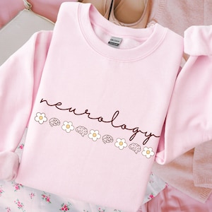 Puede incluir: Sudadera rosa claro con la palabra "neurology" escrita a mano. El dise&ntilde;o incluye peque&ntilde;as margaritas blancas e ilustraciones de cerebros. La prenda est&aacute; hecha de tela suave.