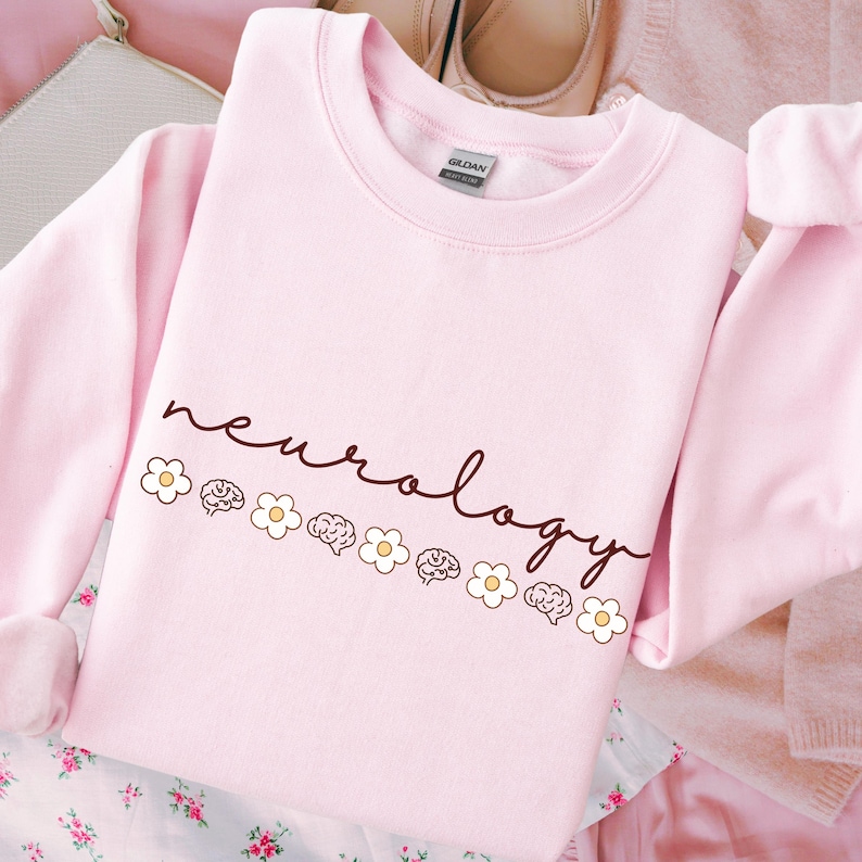 Puede incluir: Sudadera rosa claro con la palabra "neurology" escrita a mano. El dise&ntilde;o incluye peque&ntilde;as margaritas blancas e ilustraciones de cerebros. La prenda est&aacute; hecha de tela suave.