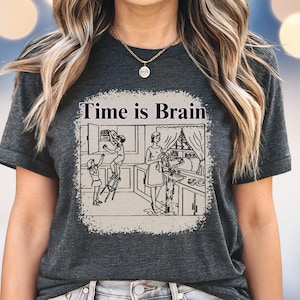 May include: Dark gray t-shirt featuring a distressed graphic with the text "Time is Brain." The graphic depicts a vintage kitchen scene with people cooking and reaching for items. The shirt has a crew neck and short sleeves.