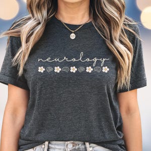 Puede incluir: Camiseta gris oscuro con la palabra "neurology" en fuente manuscrita. Debajo, se alternan margaritas blancas e ilustraciones de cerebros. La camiseta es de cuello redondo y est&aacute; hecha de un material suave.