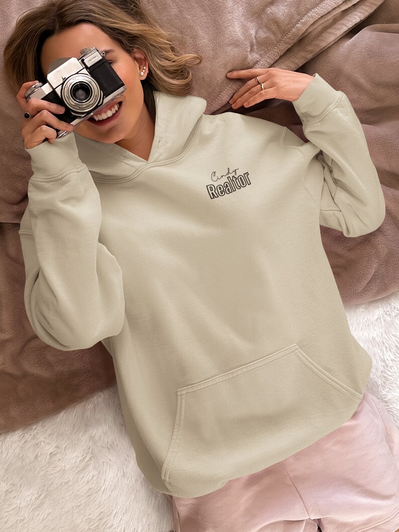 May include: A light beige hoodie with a large pocket on the front. The hoodie has the text "Cindy Realtor" printed on the front in black.