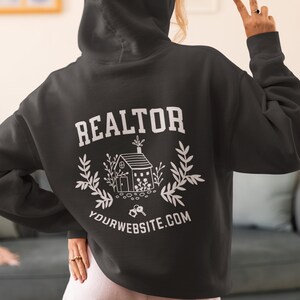 May include: Black hooded sweatshirt with the word "REALTOR" in white text on the back. A white illustration of a house with keys and greenery is below the text. The text "YOURWEBSITE.COM" is below the illustration.