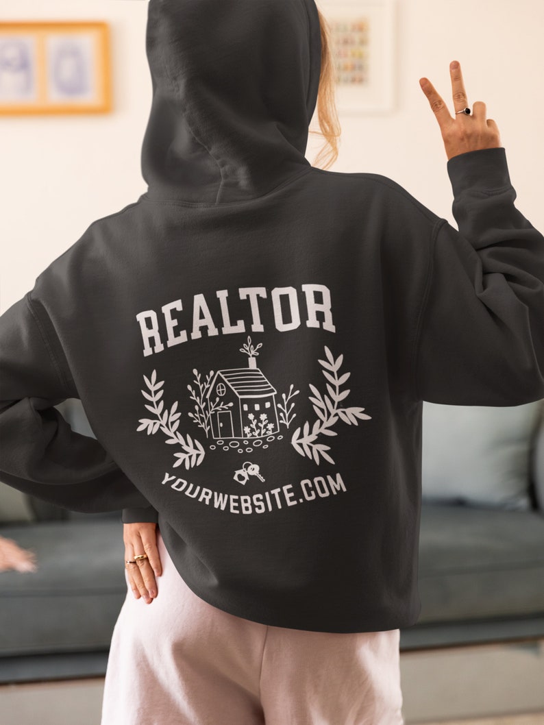 May include: Black hooded sweatshirt with the word "REALTOR" in white text on the back. A white illustration of a house with keys and greenery is below the text. The text "YOURWEBSITE.COM" is below the illustration.