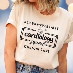May include: Cream-colored t-shirt with the text "ALL DAY EVERY DAY" above a rectangular design. Inside the rectangle is the word "cardiology squad" and a heart graphic. The words "Custom Text" are below the design.