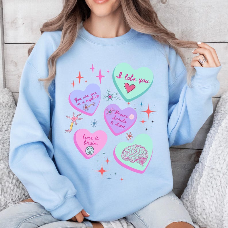 May include: Light blue sweatshirt with heart-shaped graphics and text. The hearts feature phrases like "I love you" and "Brains Scrubs & Love," along with brain illustrations and star accents. A casual, comfortable top.
